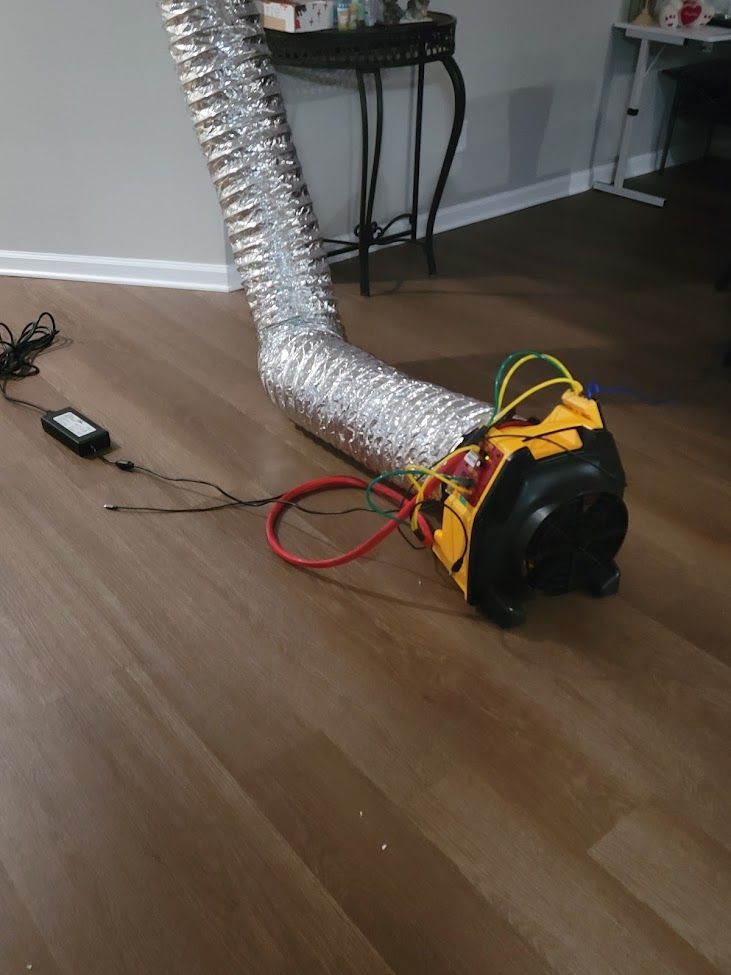 A yellow and black ventilation fan with a long, flexible silver duct tube attached, resting on a hardwood floor.