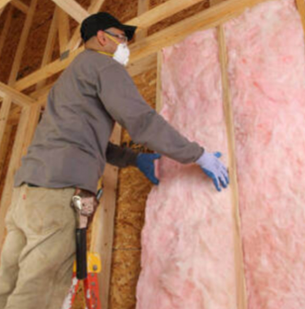 A person wearing a face mask and gloves installs pink fiberglass insulation between wooden wall studs.