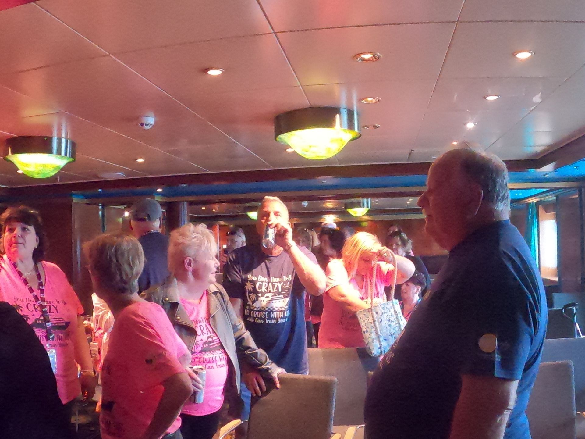 People socializing in a brightly lit bar, some wearing pink. Man in foreground looking right.