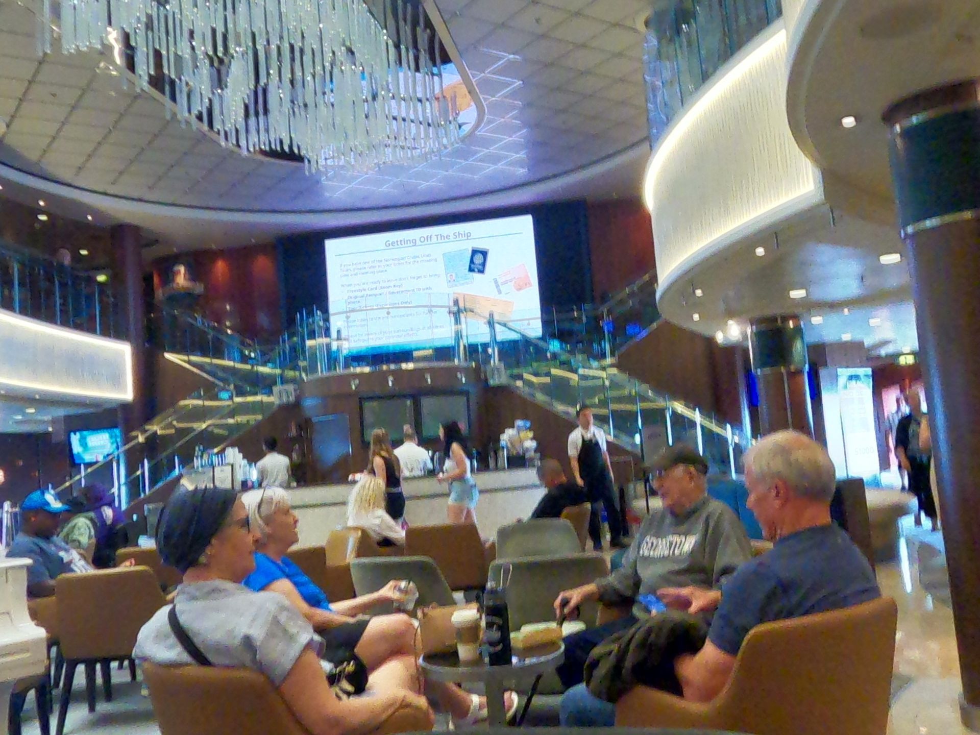 People relax in a cruise ship lobby with a large screen and a grand staircase.