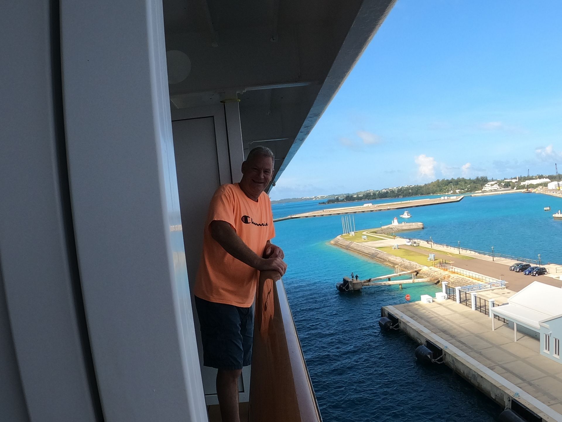 Man on cruise ship balcony, wearing orange shirt, overlooks a harbor with blue water and blue sky.