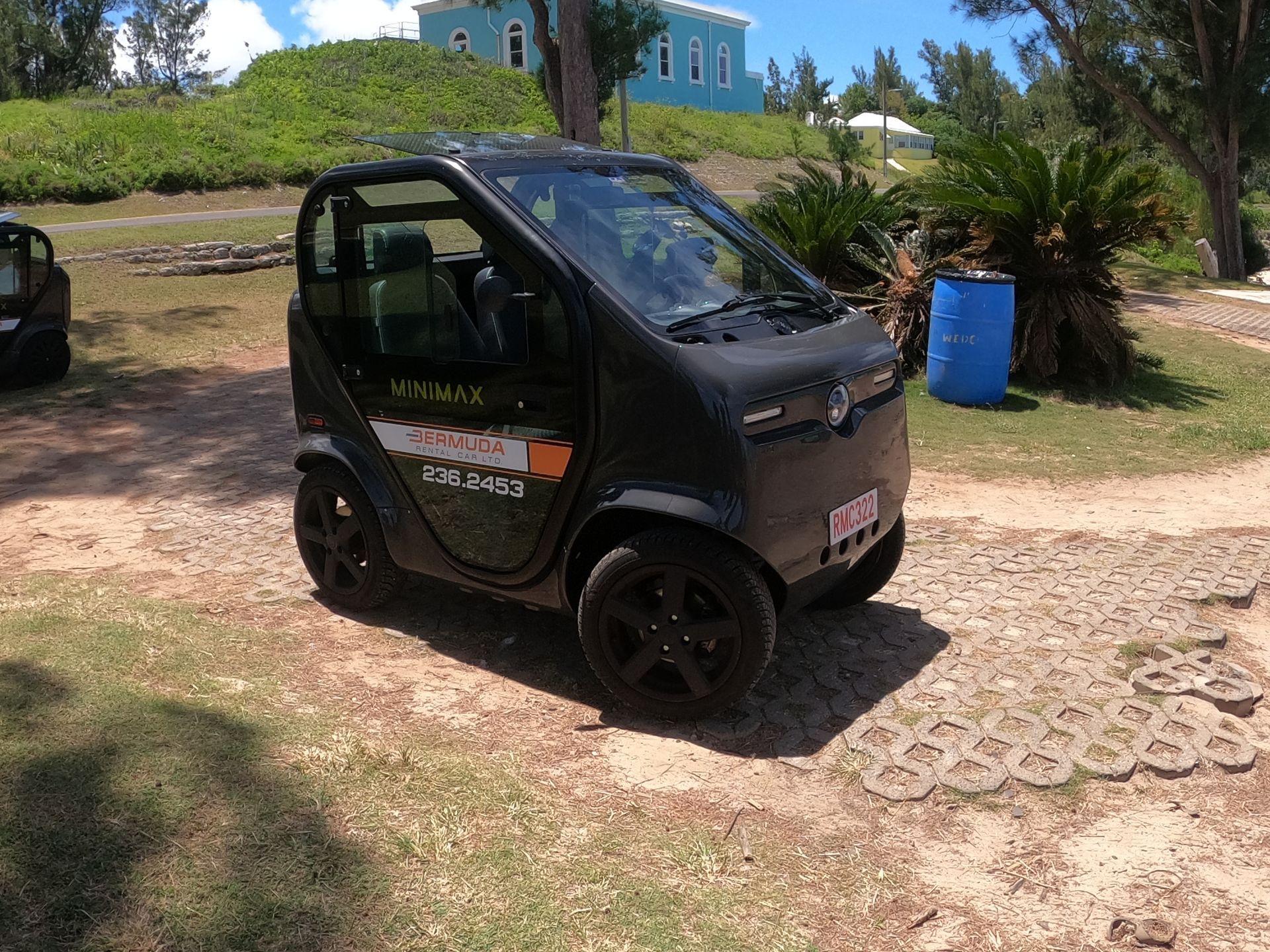 Black electric car on a sandy path near a beach with green vegetation and a blue building in the background.