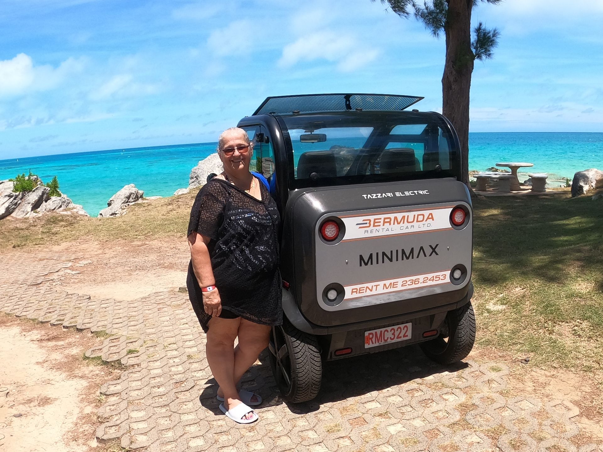 Woman posing with a small, grey rental car (MINIMAX) near a turquoise ocean and rocky shoreline.