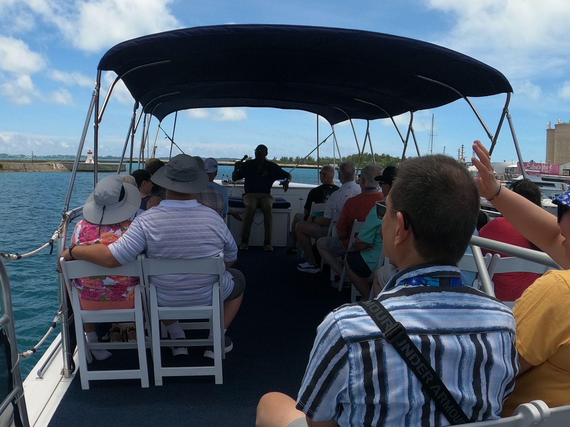 People on a boat tour under a blue canopy, with a guide and harbor in the background.