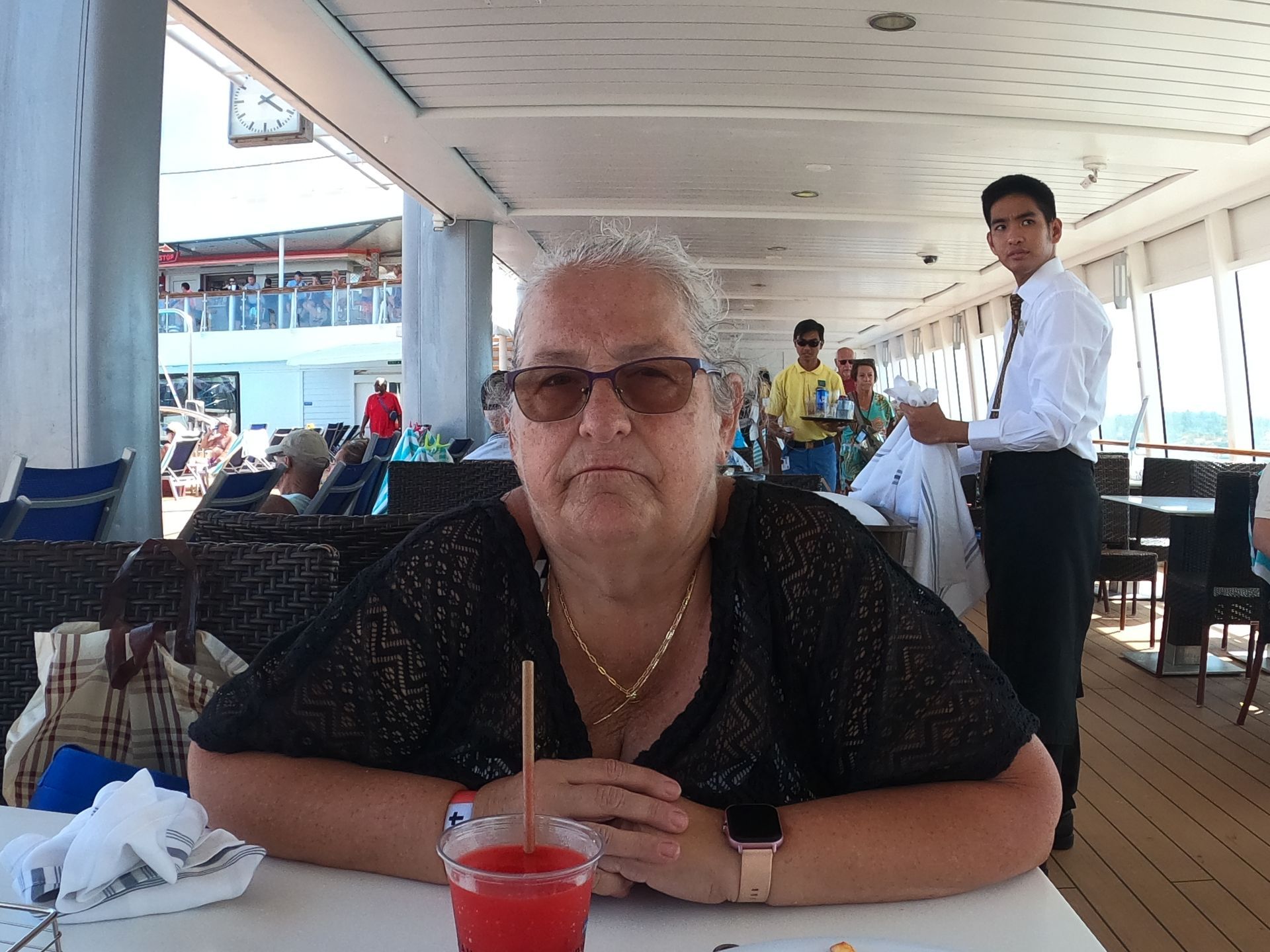 Woman with sunglasses seated at a table on a cruise deck; a waiter stands nearby.