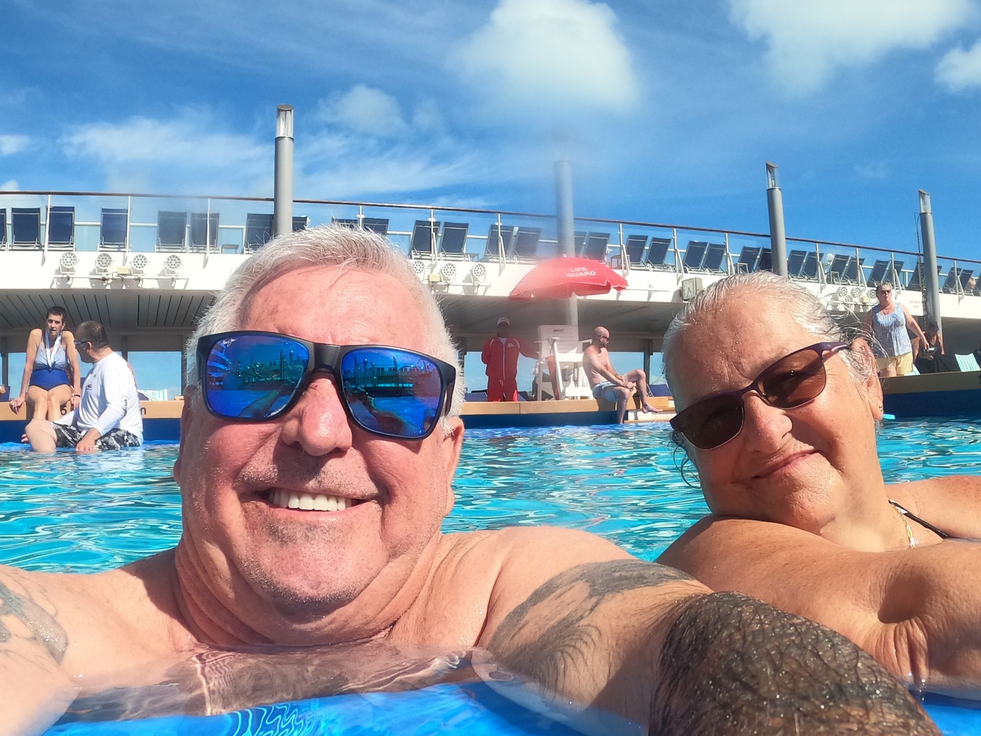 Smiling couple in a pool on a cruise ship, wearing sunglasses. Blue water and sky, sunny day.