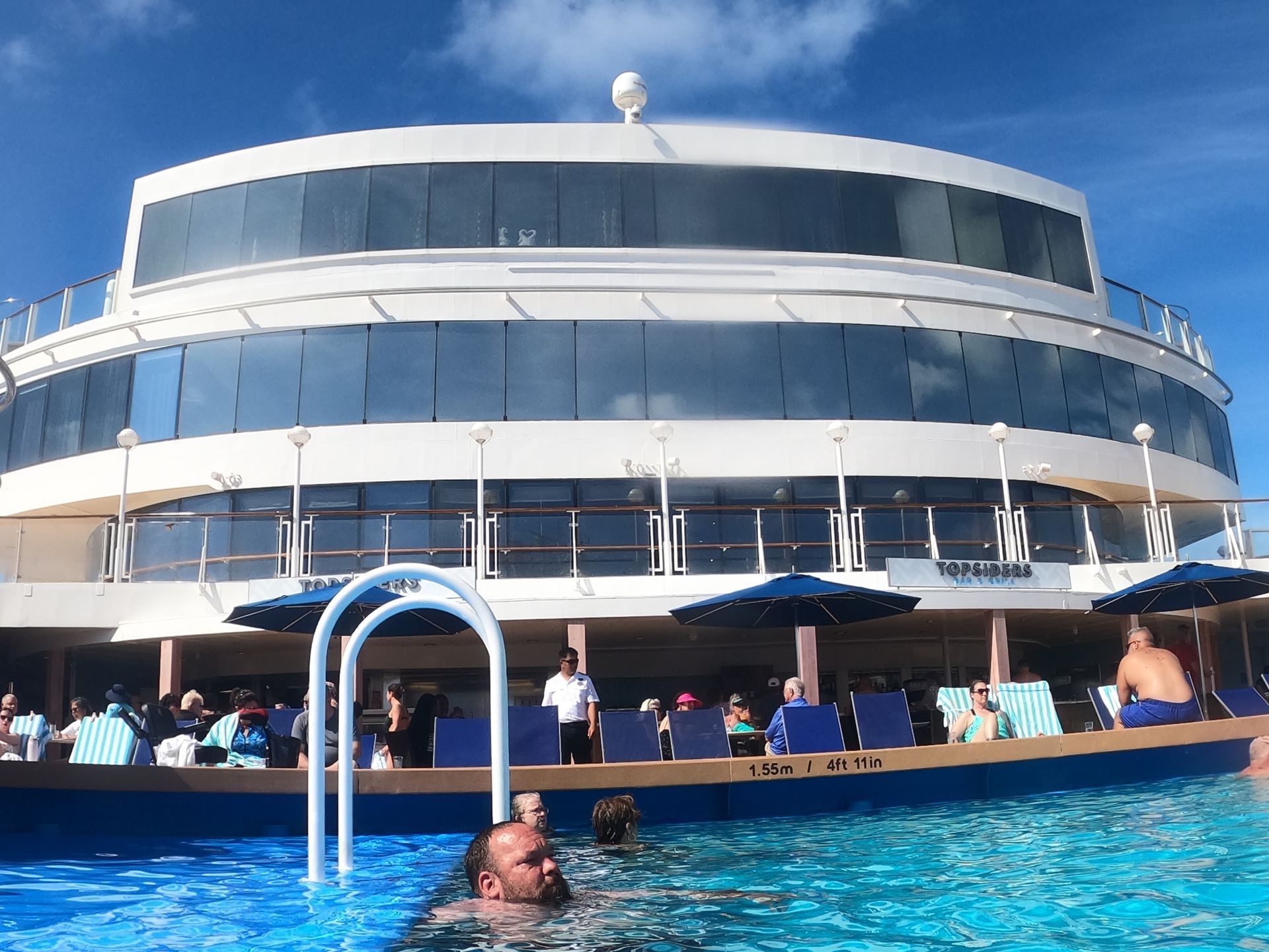 A cruise ship pool with people swimming and sunbathing, with a multi-level building in the background.