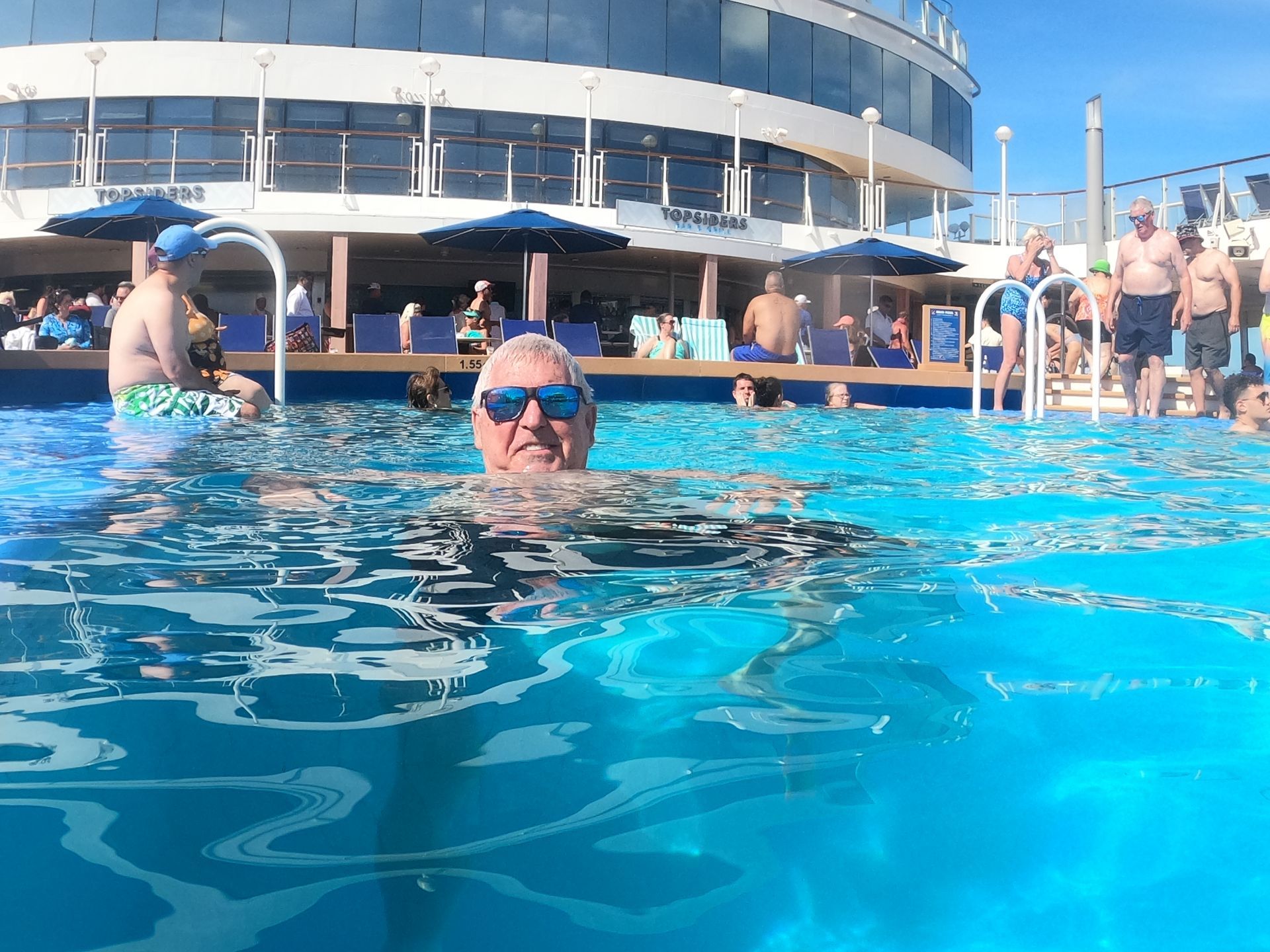 Man in sunglasses in a swimming pool on a cruise ship, sunny day. People lounging around the pool.