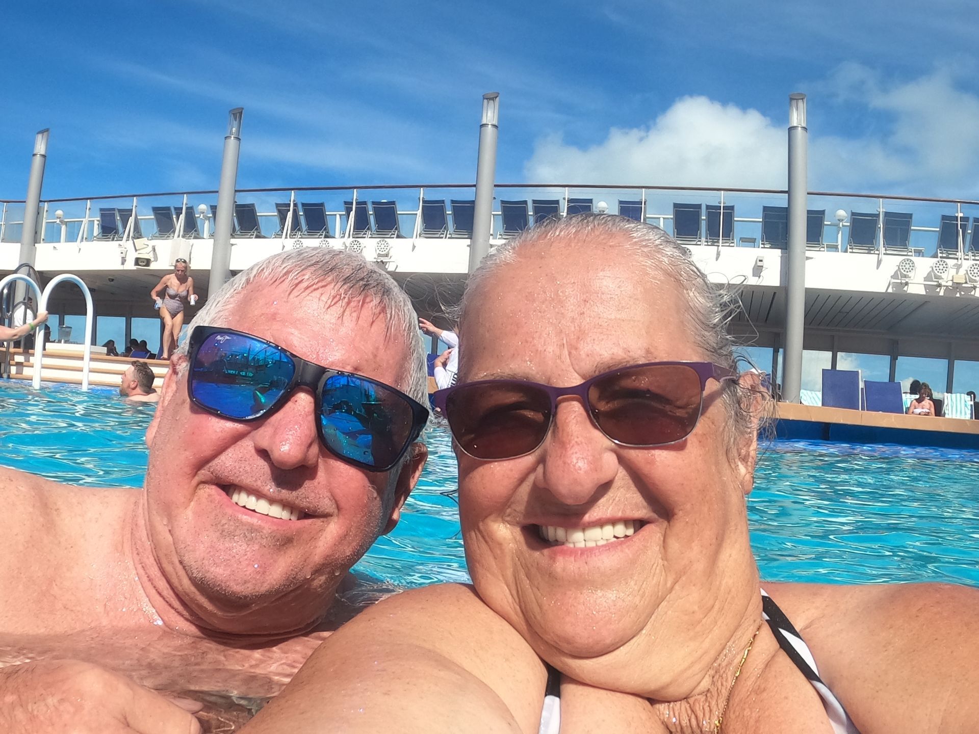 Couple in pool, smiling, wearing sunglasses. Ship deck, blue sky background.