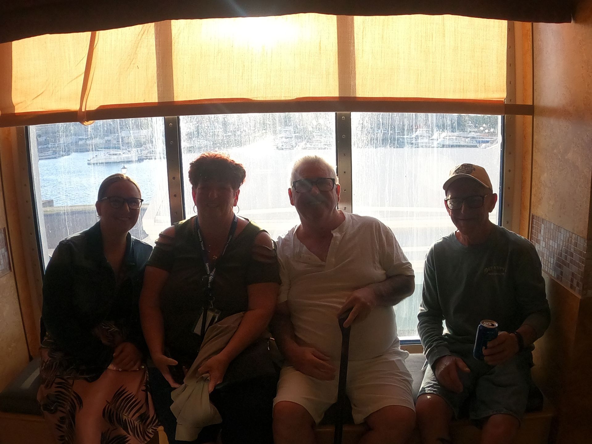 Four people seated by a window, possibly on a boat. The window shows water and land outside.