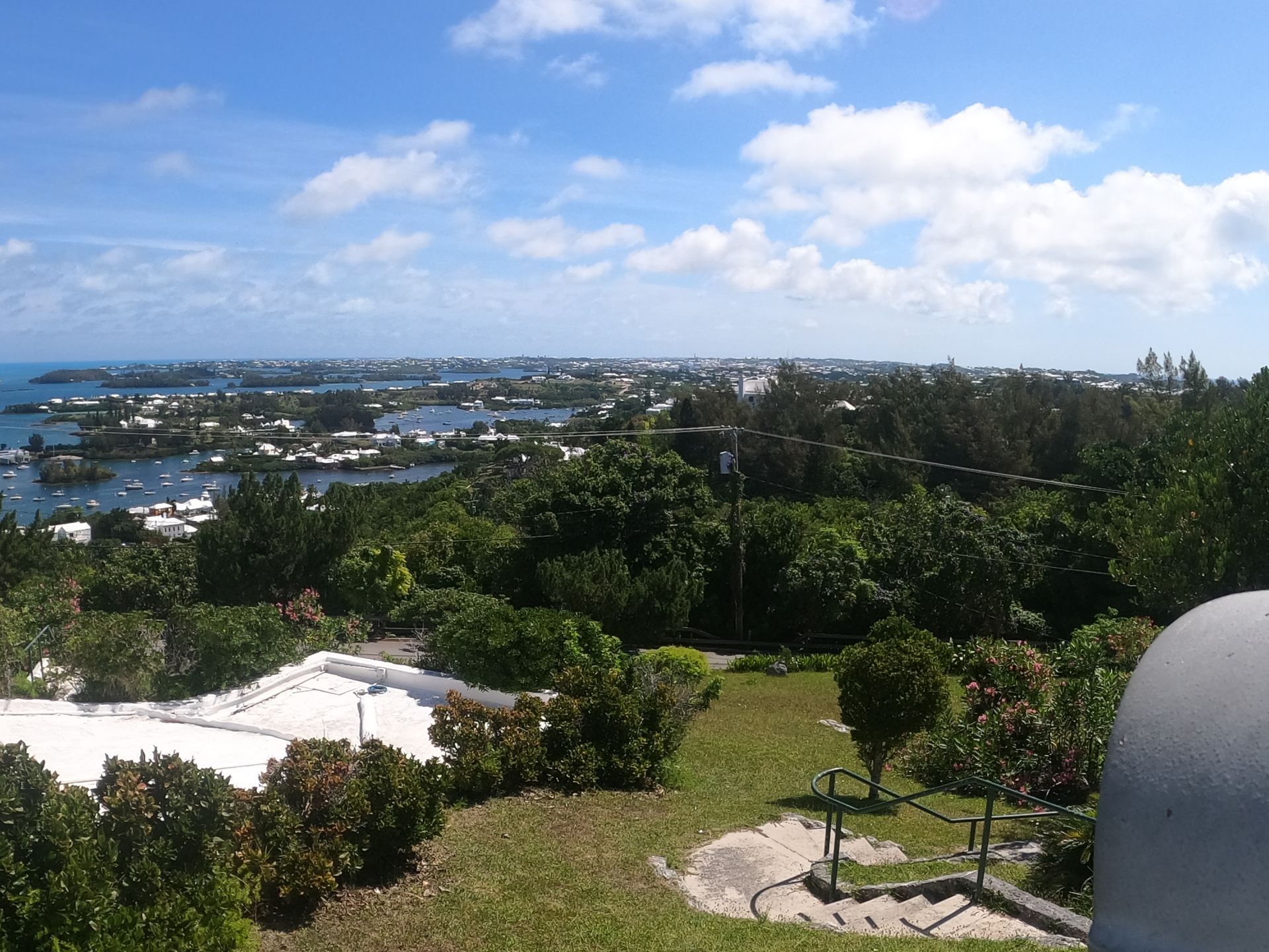 Overlooking a coastal town with boats, white rooftops, green trees, and a blue sky with clouds.