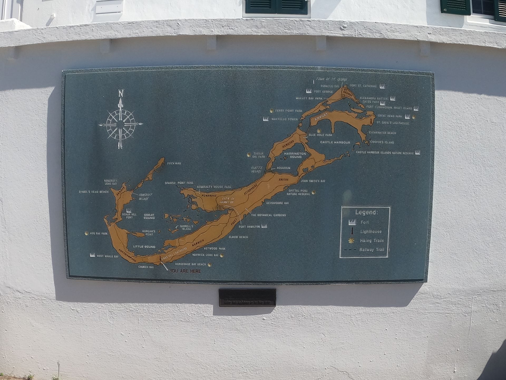 Map of Bermuda in gold against a dark grey background, mounted on a white wall, with a compass rose.