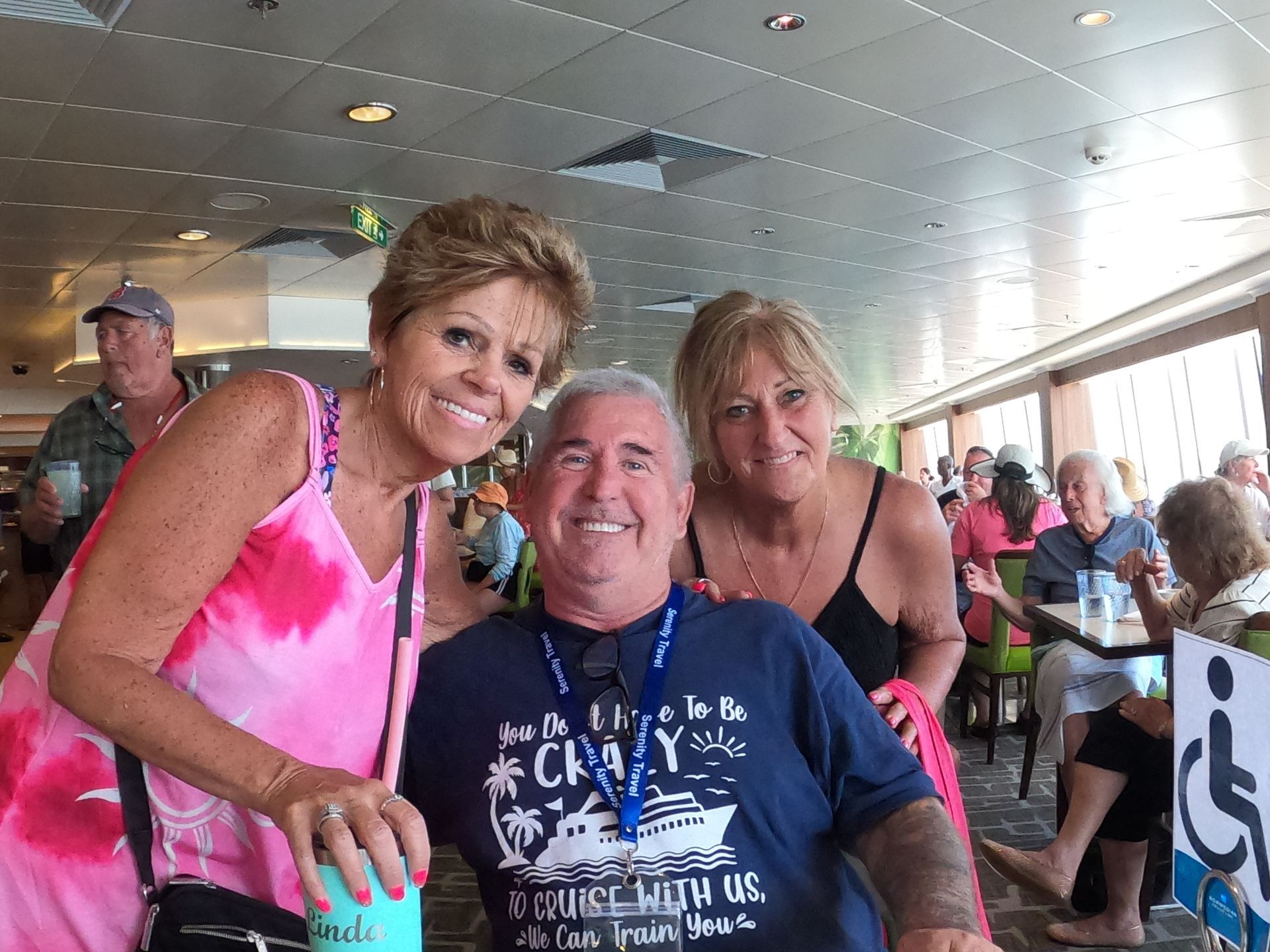 Three people smile; one in wheelchair. Inside a bright, casual dining area on a cruise ship.