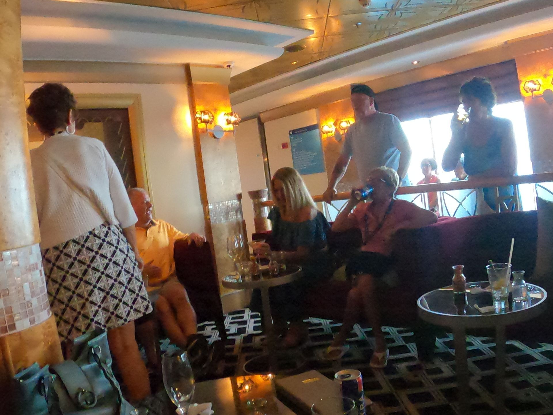 People socializing in a lounge on a cruise ship, some seated, some standing. Warm tones and cozy seating.