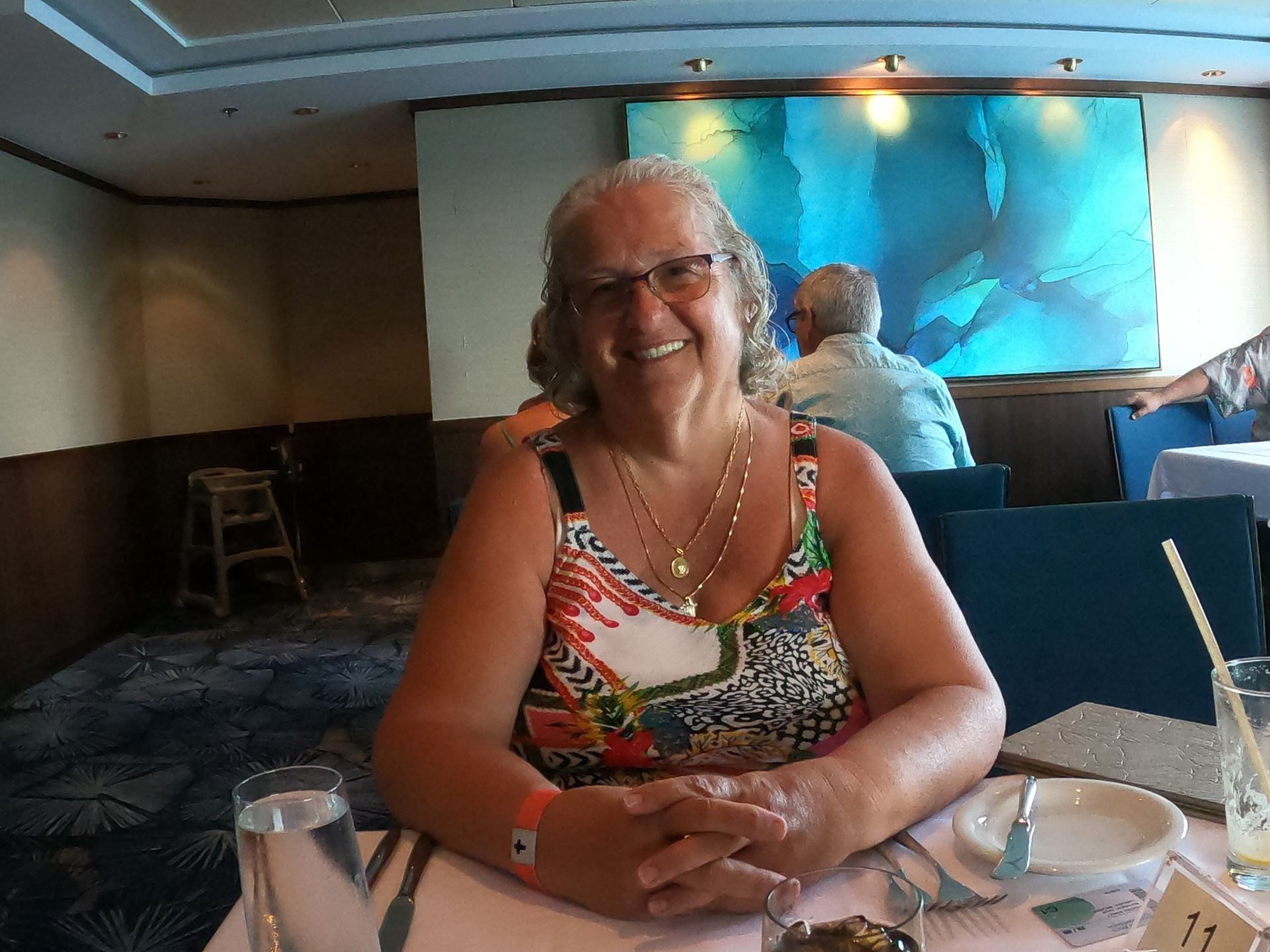 Woman with glasses smiles at a table in a restaurant; painting in background.