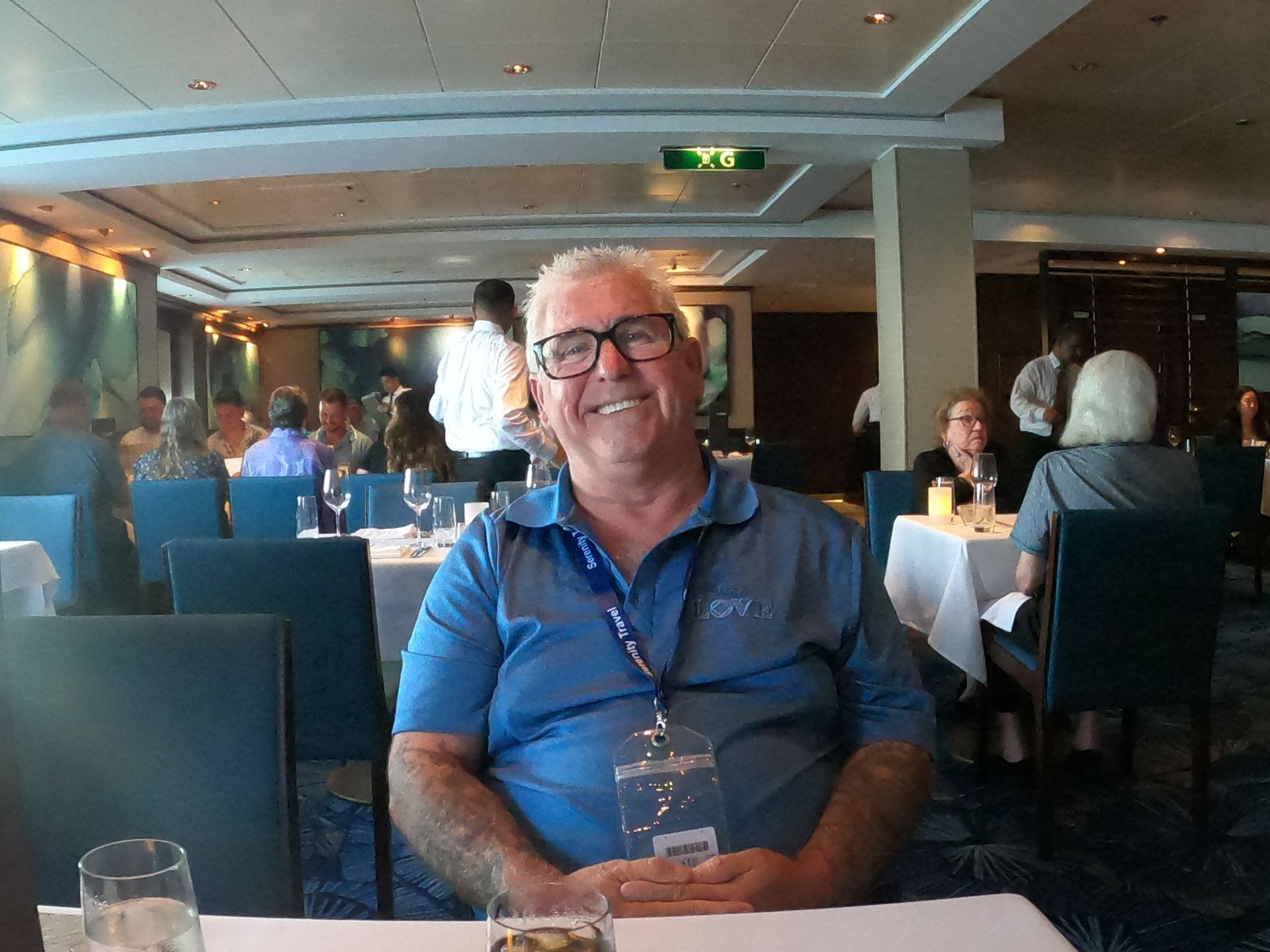 Man in blue shirt smiles at a table in a dining room; people and ocean-themed art in the background.