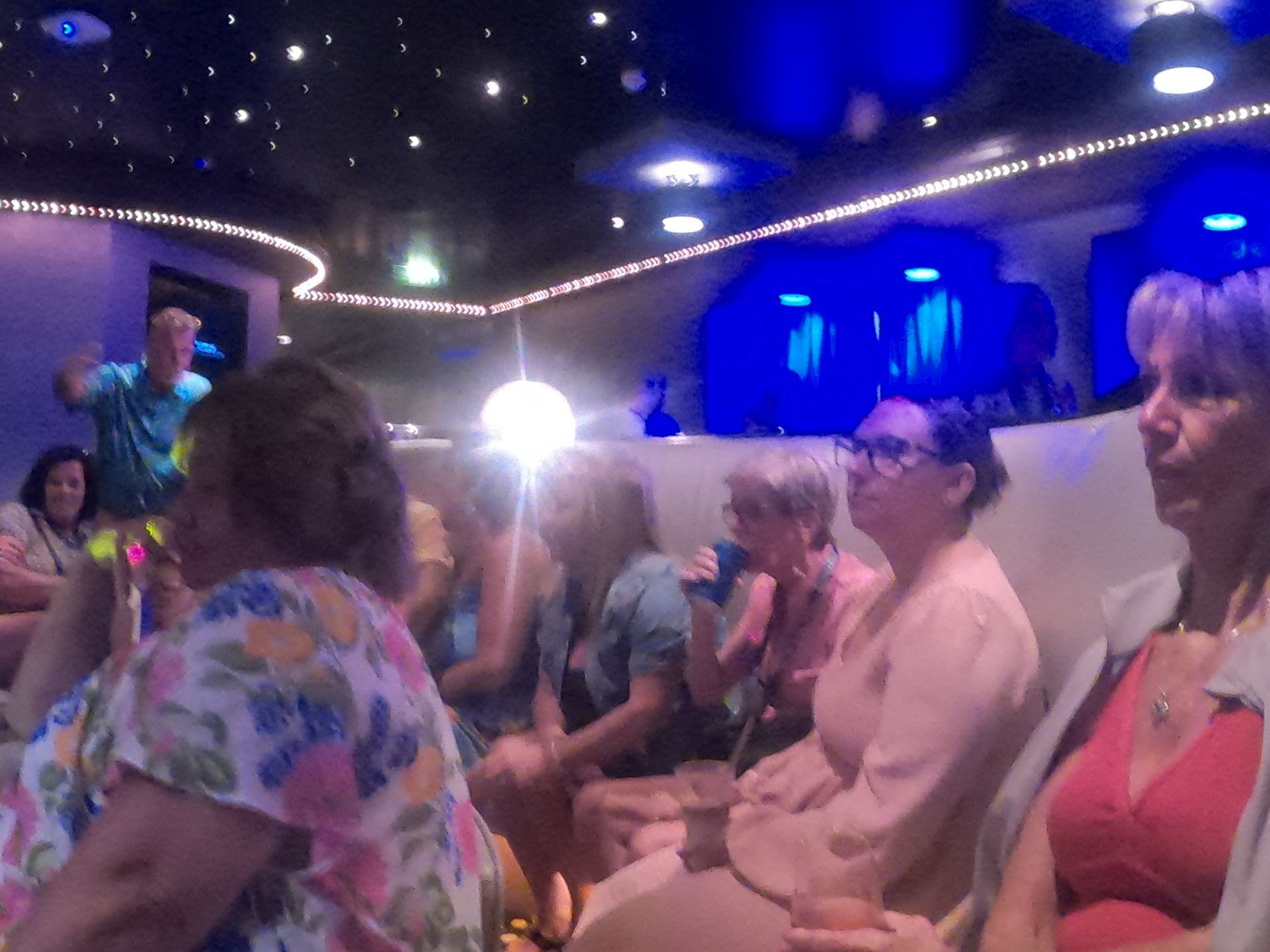 People seated in a dimly lit room with a performer. Blue and white lights, floral tops.