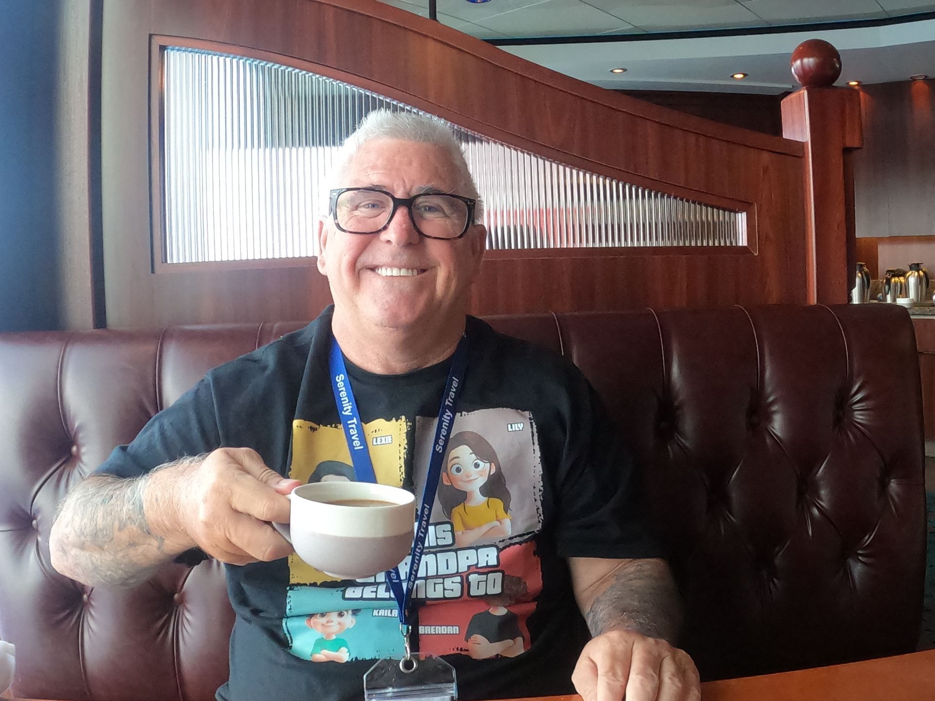 Man with glasses smiling, holding coffee cup in a booth, black t-shirt, indoor setting.