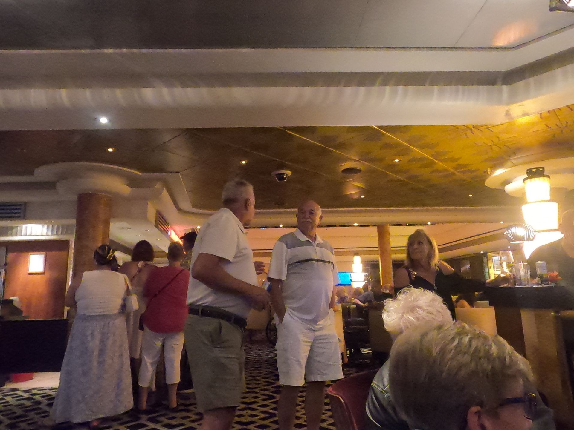 People in a dimly lit bar. Men in shorts, woman in dresses, socializing. Gold ceiling, checkered floor, warm lighting.