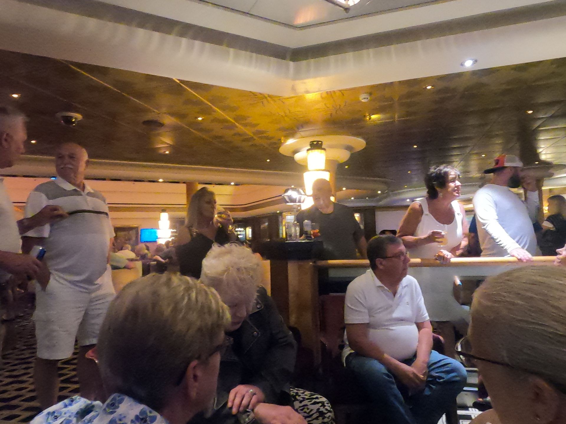 People gathered in a dimly lit bar. Some are sitting, others standing, talking and drinking.
