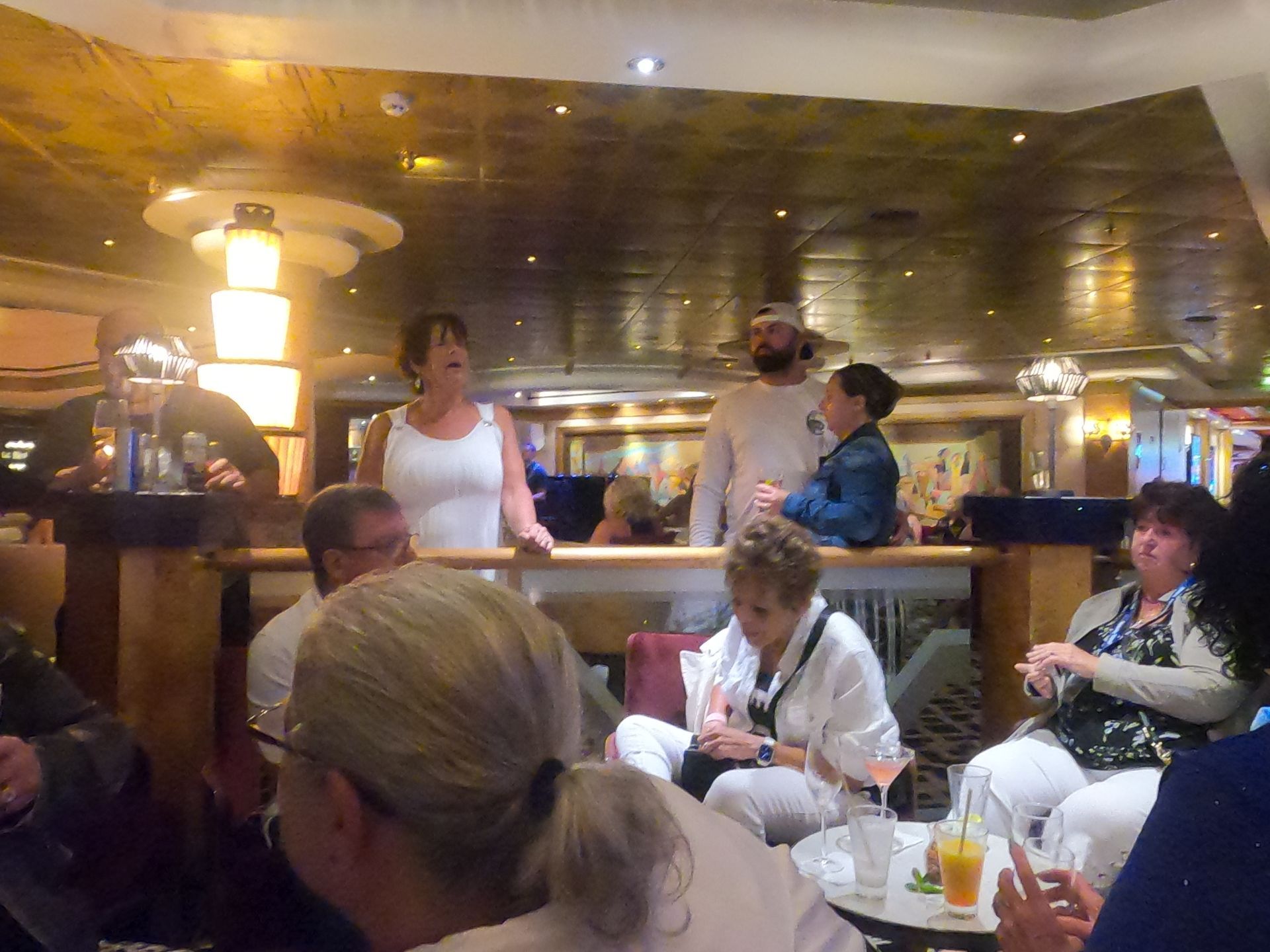 People in a bar, some standing, talking, others seated, drinking. Interior, warm lighting.