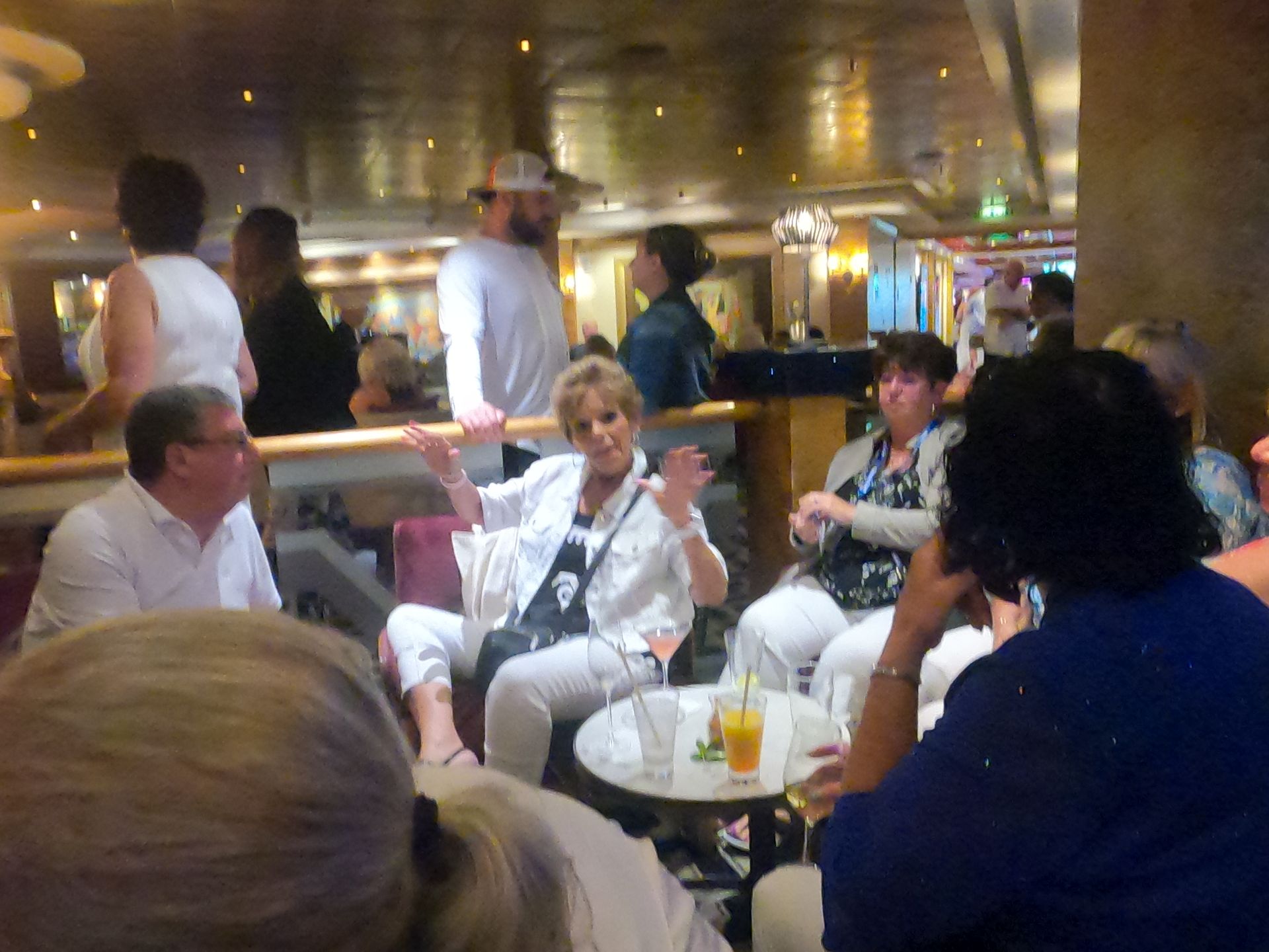 People socializing in a dimly lit lounge. Woman in white jacket gestures, others seated with drinks.