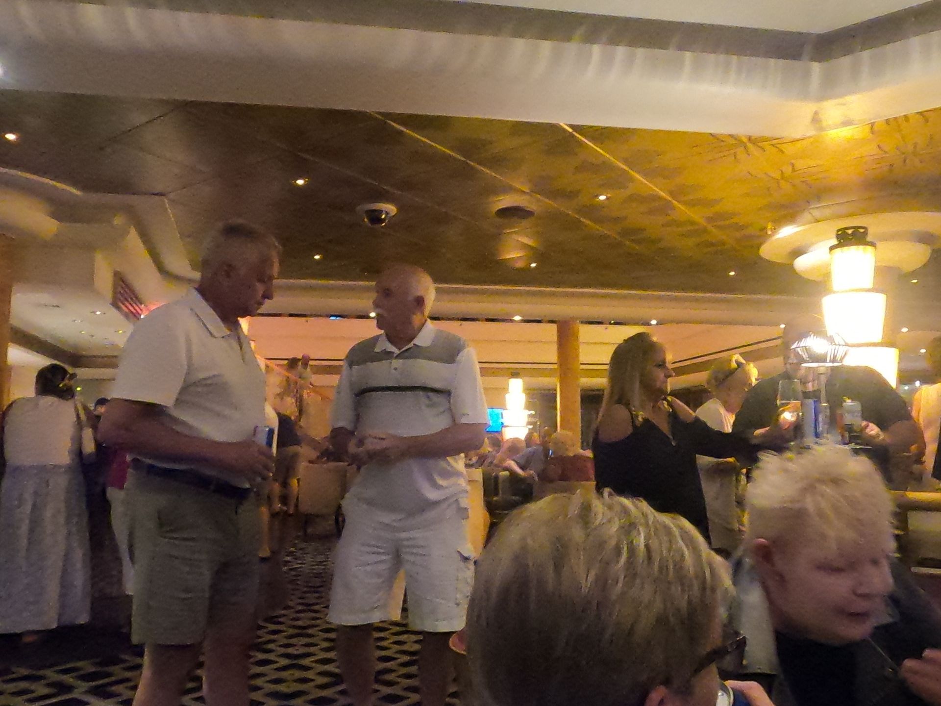 Two men talking in a brightly lit room with others, a woman taking a photo, and another person drinking.