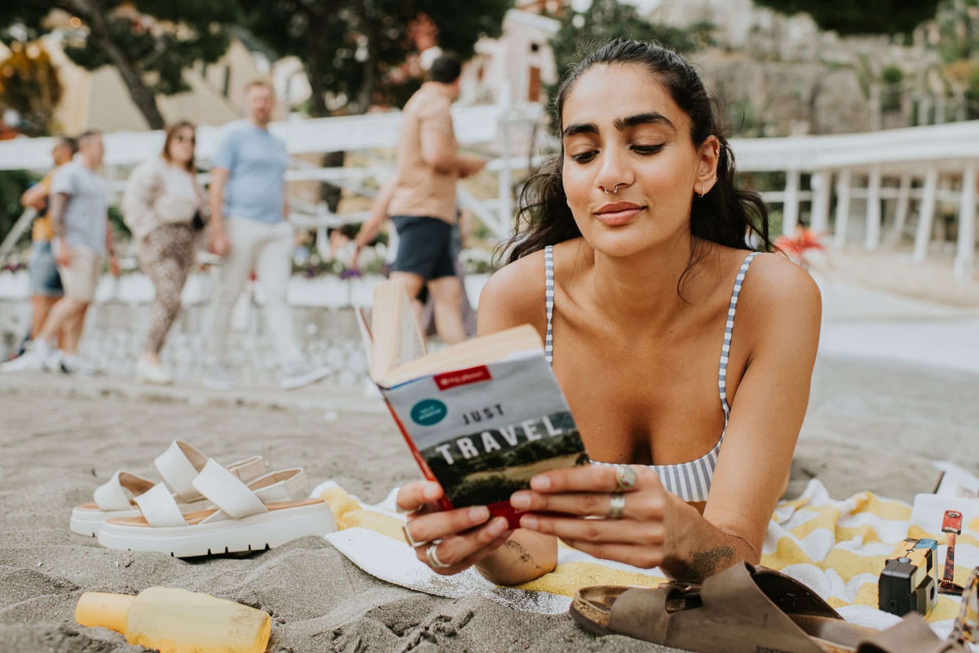 Person relaxes on beach towel reading “Just Travel” book near sandals and a bottle.