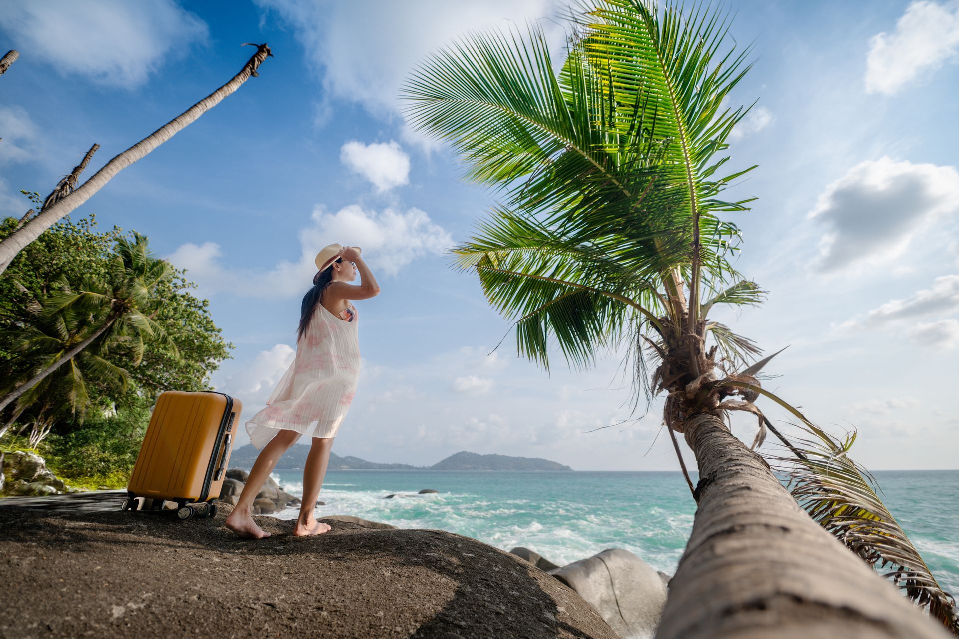 A woman in a white dress stands on the earth in front of a beach, looking around.