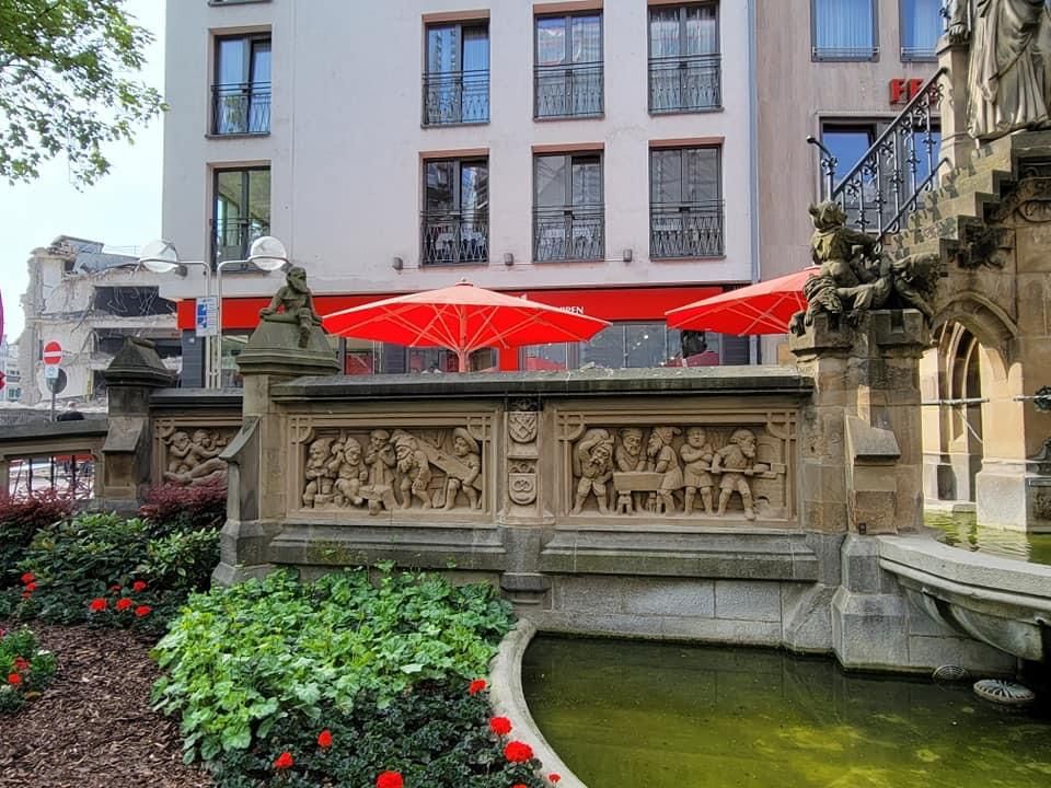 Stone fountain with carved relief, red umbrellas in front of a building with windows.
