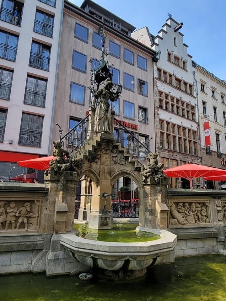 Fountain in a city square, with sculptures, water, and surrounding buildings.