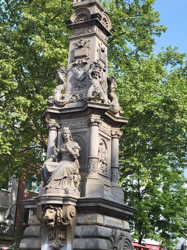 Statue of a seated queen holding a staff, with elaborate carvings and lion head fountain, in a park setting.
