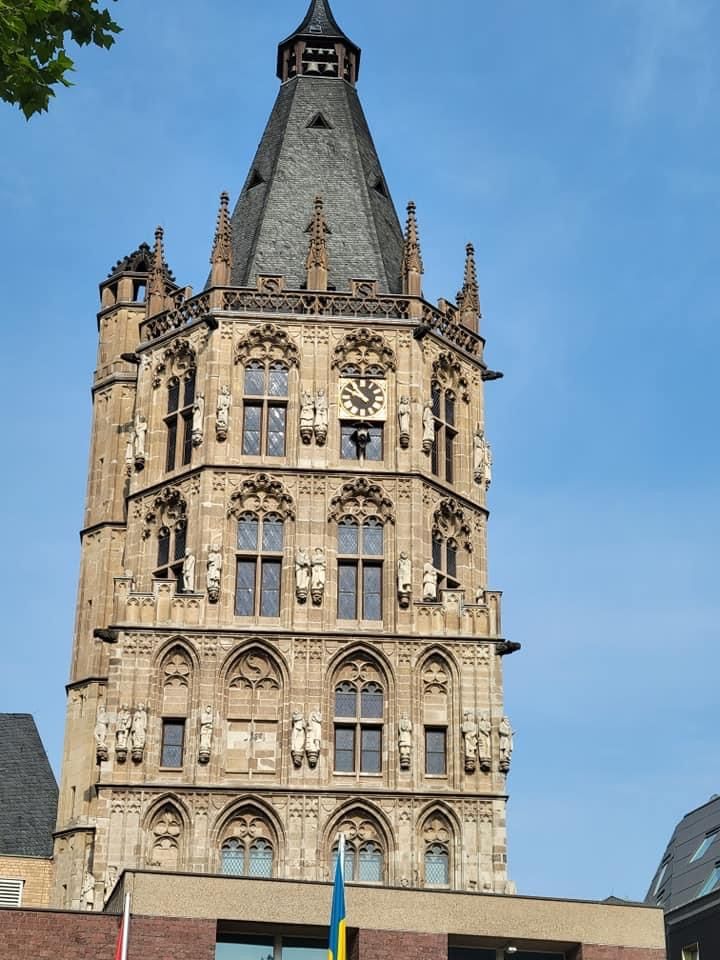 Tower of Cologne's City Hall, tall stone building with intricate carvings and clock, blue sky background.
