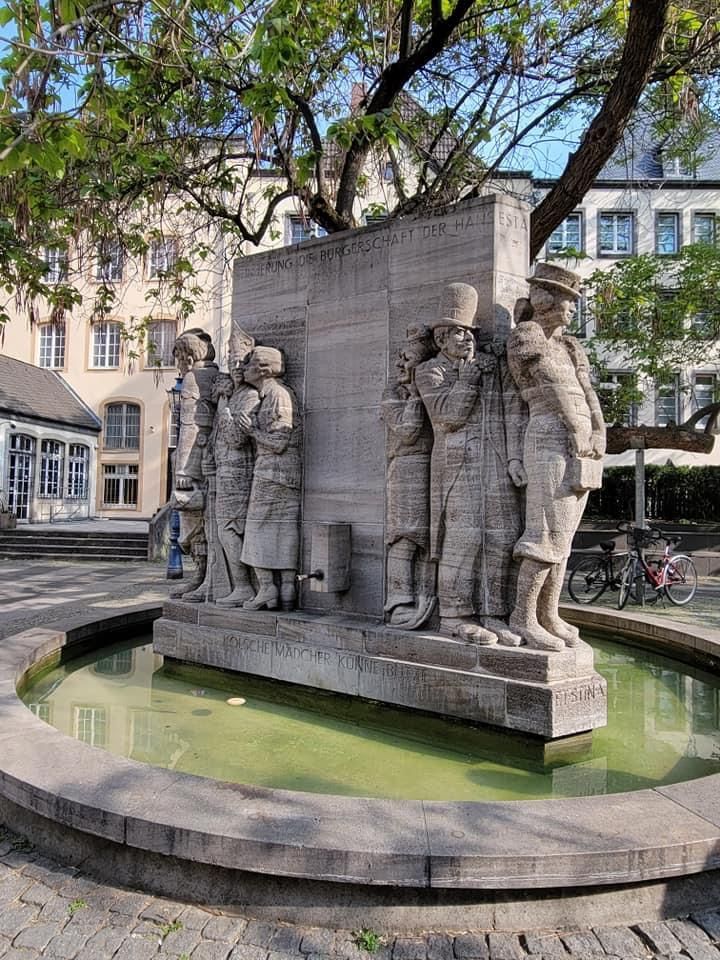Stone fountain with figures in a plaza. Green water surrounds the sculptures. Buildings and trees in the background.