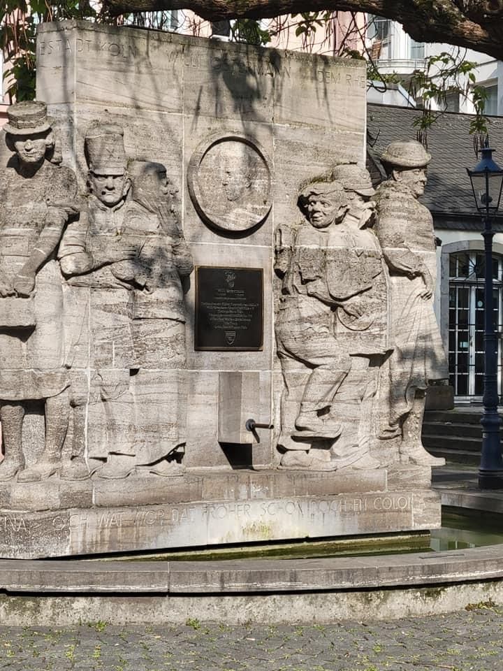 Stone sculpture in a square with figures, a portrait, and plaque, possibly a historical monument.