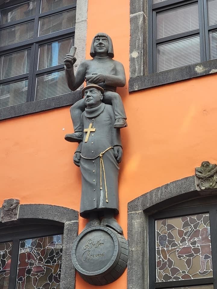 Statue of a man riding on the shoulders of a monk, mounted on a barrel, orange building.