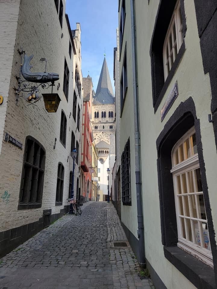 Narrow cobblestone alley, flanked by buildings, leads to a church tower in Cologne, Germany.