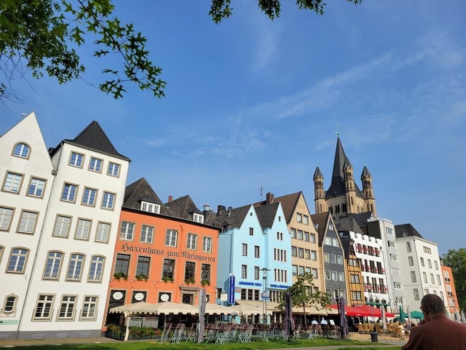 Colorful buildings along a river in Cologne, Germany, with the cathedral in the background and a clear blue sky.