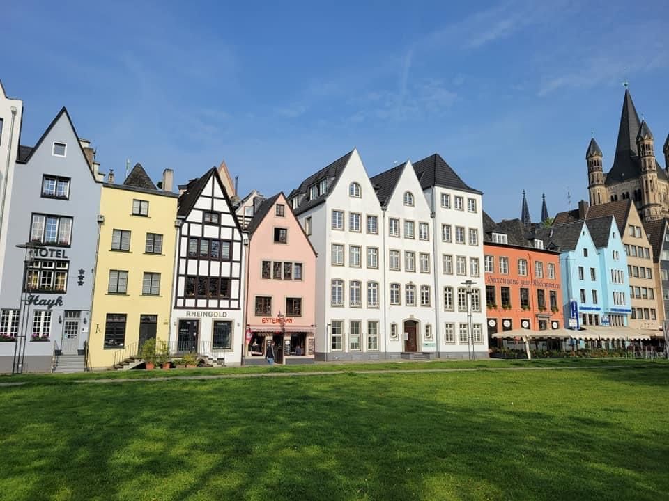 Colorful buildings line a green lawn with the Cologne Cathedral in the background, under a blue sky.
