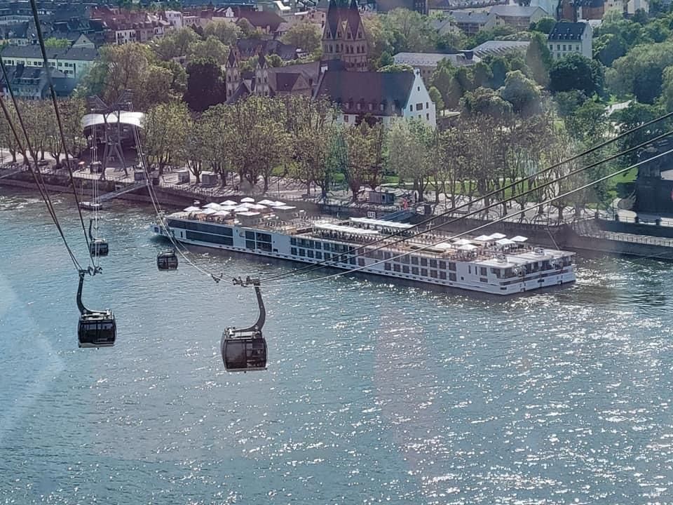 Gondola cable cars over river; large white cruise ship docked; buildings and trees in background.