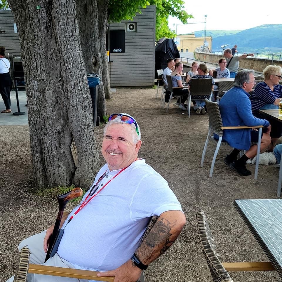 Man smiling at an outdoor cafe, holding a cane. Other people seated at tables.