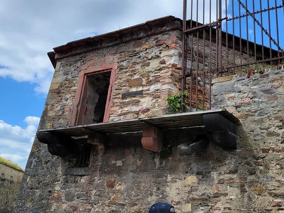 Stone turret with an open doorway, metal bars, and a small balcony, against a cloudy sky.