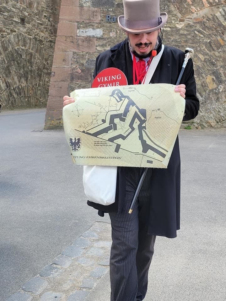 Man in top hat holding a map with a Viking illustration. Outdoors, near stone wall.