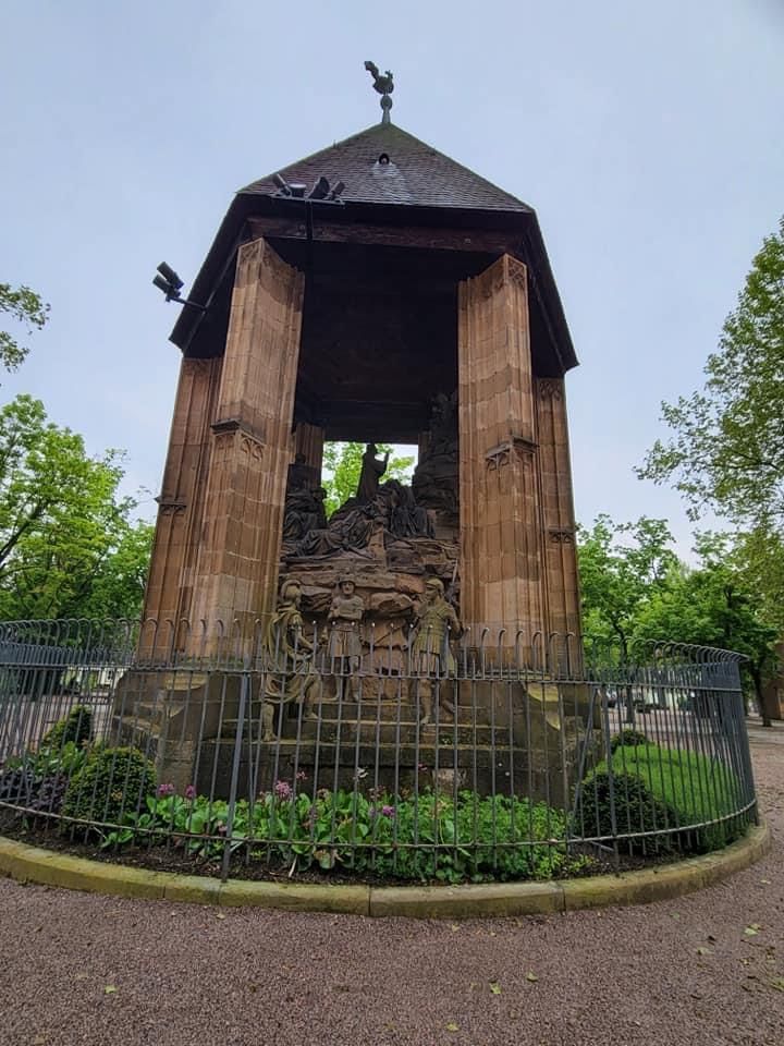 Stone monument with statue under a dark roof, fenced in with flowers in front.