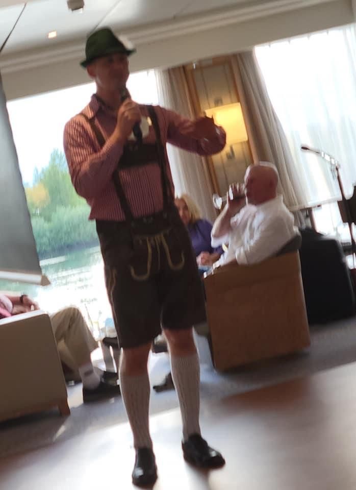Man in lederhosen, holding a microphone, speaking in an indoor setting. Other people are present.
