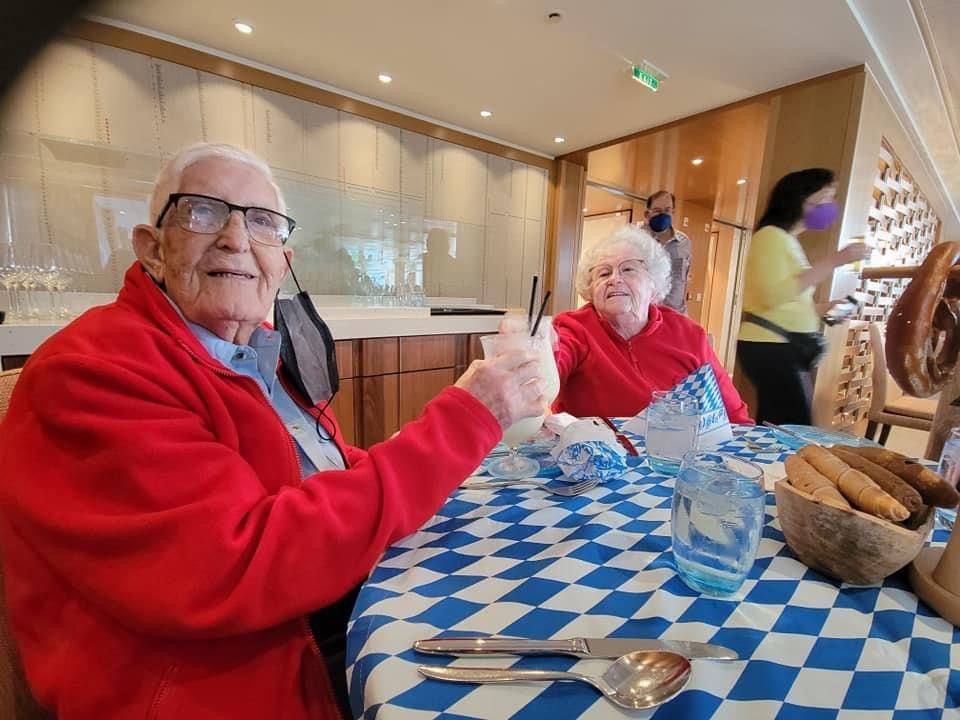 An elderly man in a red jacket gives a thumbs up at a table with a woman in a red shirt. Oktoberfest themed.