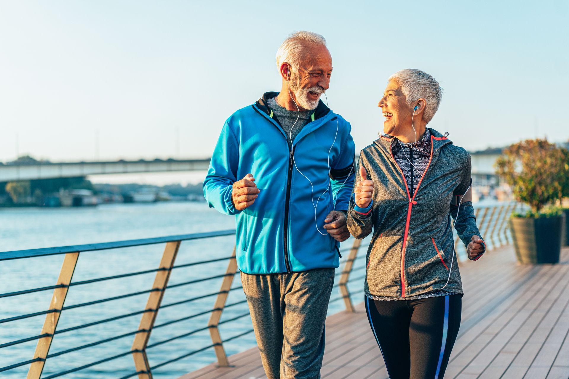 Two people jogging outdoors, smiling. Waterfront setting with blue sky.