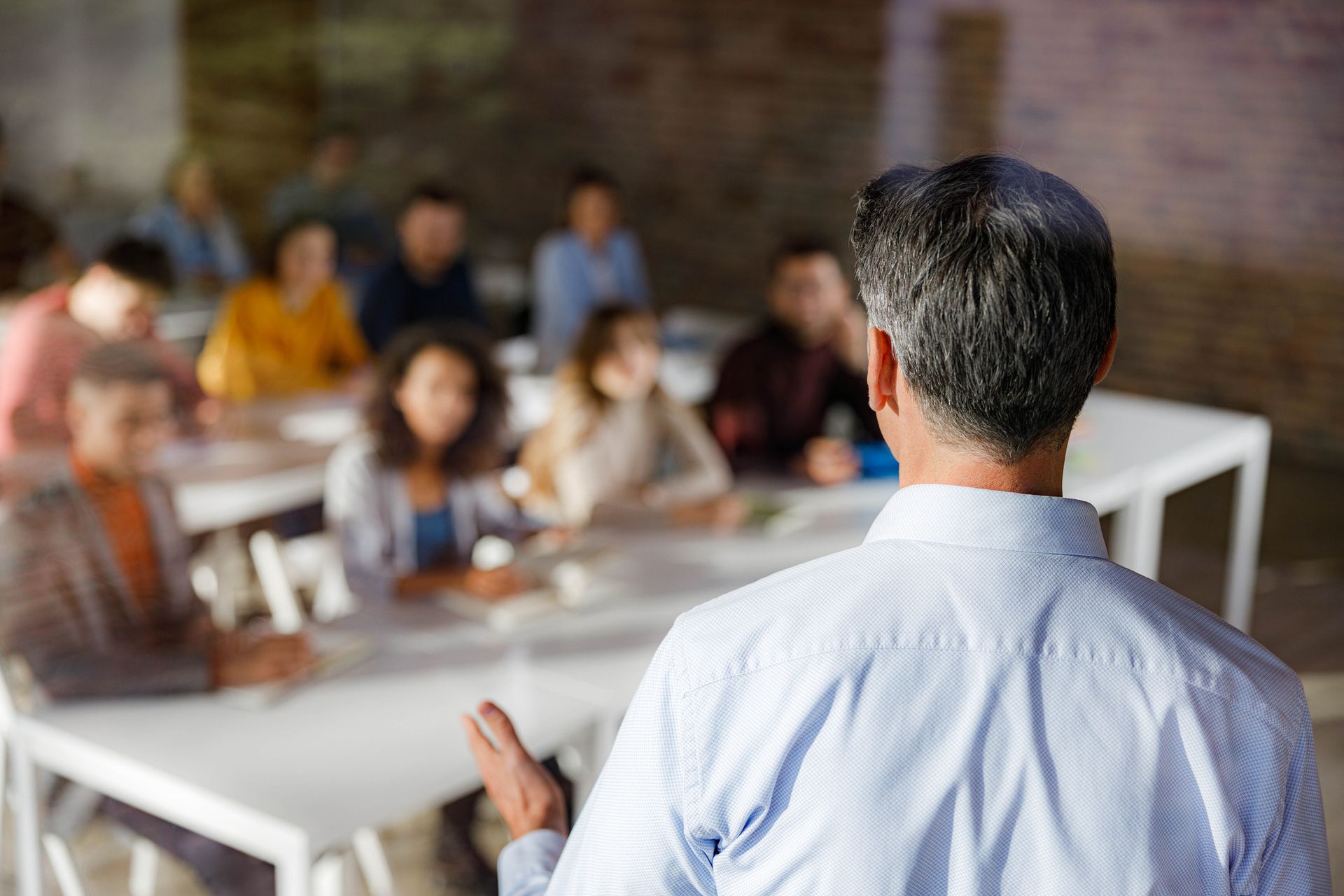 Man addressing a group of people seated at tables in a room with a brick wall background.