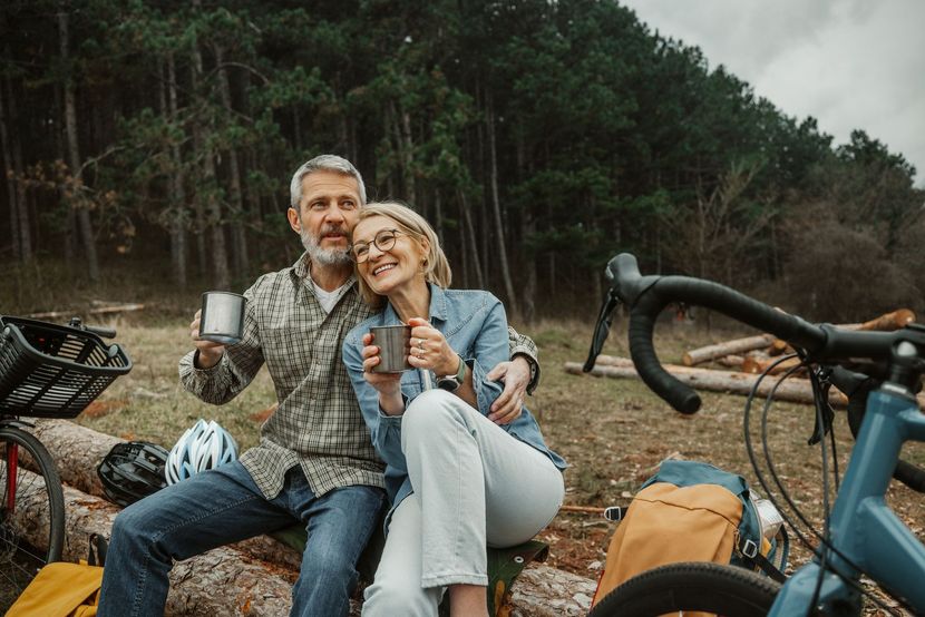 Couple on a log, holding mugs, smiling. Bicycles and forest background.
