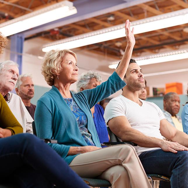 Woman raising hand, in audience at a meeting. Others seated, attentive. Indoors, bright.